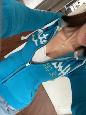 Blå tajt zip up hoodie från Hollister - Snygg blå hoodie från Hollister med vit broderad logga och text på bröstet och armen