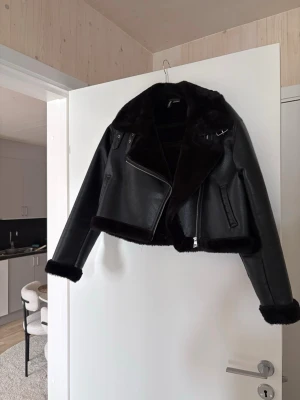 Svart biker skinnjacka med fuskpäls - Snygg svart biker skinnjacka från H&M med mjuk fuskpäls på krage, ärmslut och nederkant. Jackan har silvriga dragkedjor och spänndetaljer vid kragen. Kort modell som ger en cool och trendig look.