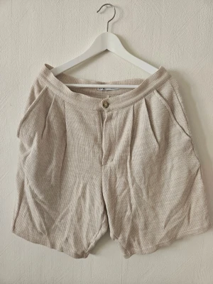 Beige shorts från Zara i bomull - Beiga shorts från Zara i mjukt bomullsmaterial med diskret struktur. Modellen är högmidjad med veck framtill, sidfickor och stängs med knapp och dragkedja. Perfekta för varma dagar och enkel att matcha med olika toppar.