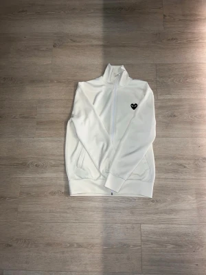 Full zip från Comme des Garçons - Säljer en vit hoodie med dragkedja från Comme des Garçons. Framtill finns ett litet svart hjärta med ögon och på ryggen ett stort svart hjärta med ögon. Hög krage och ribbade muddar. Perfekt för en stilren och trendig look.
