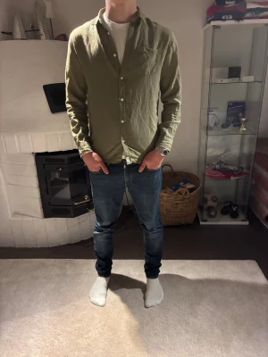 Olivgrön linneskjorta med knappar från KappAhl - Snygg olivgrön skjorta i linne med klassisk krage och knäppning framtill. Skjortan har lång ärm och en avslappnad passform, perfekt att bära över en t-shirt för en chill look. Skjortan är i gott skick: 9/10. 