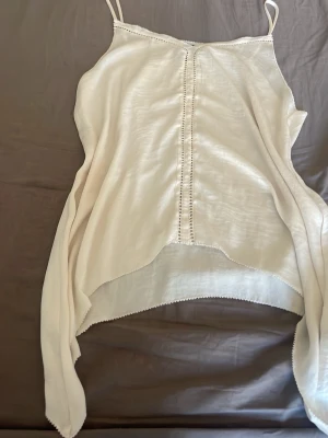 Vit linnetopp från H&M med spetsdetaljer - Säljer en vit linnetopp från H&M med tunna axelband och dekorativ spets längs kanterna och mittsömmen. Toppen har en luftig passform och är perfekt för varma dagar. Enkel och stilren design som är lätt att matcha.