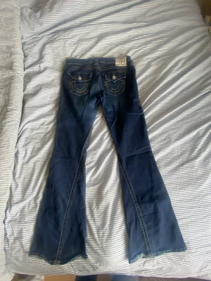 Mörkblå bootcut jeans True Religion - Säljer ett par mörkblå bootcut jeans från True Religion med snygga kontrastsömmar och detaljerade bakfickor med lock och knappar. Klassisk femficksmodell i stretchigt denimtyg. Perfekta för dig som gillar retrovibbar och markerad siluett.