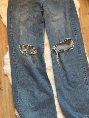 Blå slitna jeans med hål vid knäna - Säljer ett par blå jeans med raka ben och stora slitna hål vid båda knäna. Jeansen har en klassisk femficksmodell och är gjorda i kraftig denim. Perfekta för en avslappnad och trendig streetstil.