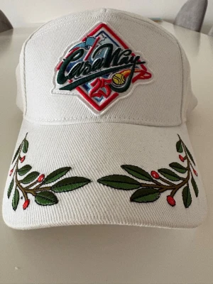 Vit keps från Casablanca Paris - Säljer en vit keps från Casablanca Paris med broderad logga och färgglada detaljer framtill. Skärmen har broderade gröna blad och röda bär som ger en unik look. Kepsen är tillverkad i tyg och har justerbar passform.