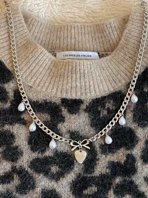 Silverfärgat halsband med pärlor och hjärta - Snyggt silverfärgat rostfritt stål halsband med kedja, dekorerat med små vita pärlor och ett hjärthänge med rosett i mitten. Perfekt accessoar för att lyfta din outfit med en söt och trendig touch. Passar dig som gillar detaljer och vill sticka ut lite extra.