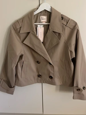 Kort beige trenchcoat från Gina Tricot - Stilren kort trenchcoat i beige från Gina Tricot. Den är i storlek 158/164. Helt ny med prislapp