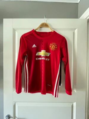 Manchester United långärmad tröja Martial - Röd långärmad Manchester United matchtröja från Adidas med Martial och nummer 11 på ryggen. Tröjan har vita ränder på sidorna, klubbmärke på bröstet och sponsorlogga framtill. Perfekt för fotbollsfans som vill visa sitt stöd.