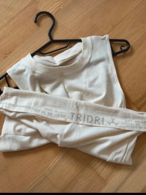 Vit ärmlös träningstopp från TriDri - Snygg och luftig vit ärmlös träningstopp från TriDri. Tillverkad i ett lätt och stretchigt material av polyamid, perfekt för intensiva träningspass. Rund halsringning och diskret meshstruktur för extra ventilation.