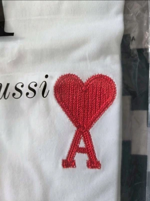 Vit t-shirt från Ami med rött hjärta - Vit t-shirt från Ami Alexandre Mattiussi med broderat rött hjärta och bokstaven A på bröstet. Klassisk rund halsringning och tryckt text framtill. Tillverkad i mjuk bomull för en skön känsla.