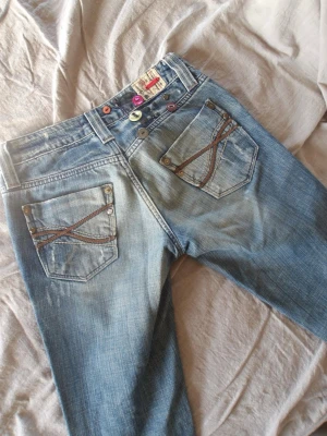 Lågmidjade jeans - Säljer mina Lågmidjade jeans med unika detaljer, de har knappar på baksidan och fina detaljer på bakfickorna.  Endast använda en gång eftersom storleken inte passar mig.  De är något lite slitna längst ner. Skriv om du önskar fler bilder på jeansen!💗 