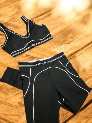 Svart träningsset från Alo - Snyggt svart träningsset från Alo med sport-bh och tights. Setet har vita kontrastsömmar som ger en sportig look. Materialet är stretchigt och passar perfekt för gym eller yoga. Både bh och tights har Alo-logga framtill.