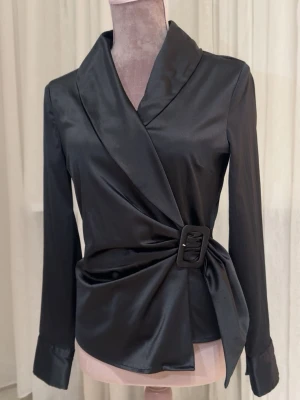 Svart omlottblus med bälte - Zara svart omlottblus i satintyg med markerad krage och lång ärm. Blusen har ett brett bälte med spänne i midjan. Storlek xs/s 