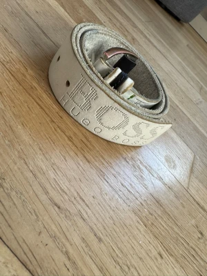 Vitt skärp från Hugo Boss i skinn - Snyggt vitt skärp från Hugo Boss i skinn med präglad logga längs hela bandet. Klassisk metallspänne och flera hål för justering. Perfekt accessoar för att lyfta din outfit med en clean och stilren look.