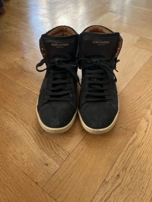 Saint Laurent svarta mocka sneakers - Säljer ett par svarta high-top sneakers från Saint Laurent i mocka med coola fransdetaljer baktill. Skorna har vita sulor, svarta skosnören och bruna detaljer på insidan. Märkeslogga på plösen och snygg streetstil. Made in Italy 
