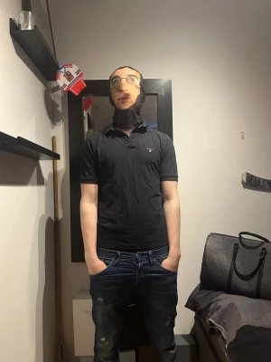 Svart pikétröja från GANT - Klassisk svart pikétröja från GANT med liten logga på bröstet. Tröjan har krage, knappar vid halsen och korta ärmar. Perfekt för en clean och stilren look. Modellen är 182 och väger 75💥