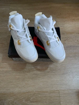 Air Jordan 4 Retro White & Gold - Snygg  Retro sneakers i helvitt läder med detaljer i guld på snörningen. Klassisk siluett med meshpaneler och synlig Air-enhet i sulan. Perfekta för dig som vill sticka ut med stilrena och exklusiva sneakers.