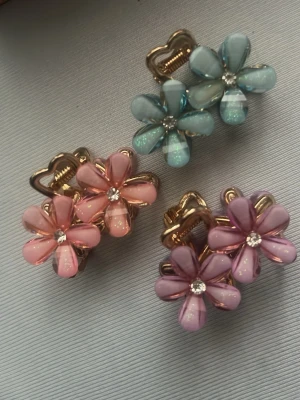 Blommiga hårklämmor i pastell - Säljer tre söta hårklämmor i form av blommor med pastellfärgade kronblad i blått, rosa och lila. Varje blomma har en glittrande sten i mitten och guldfärgad metallram. Perfekt accessoar för att piffa upp frisyren med en färgklick.