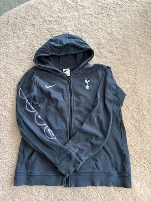 Tottenham Nike hoodie med dragkedja - Mörkblå hoodie från Nike med Tottenham Hotspur-logga på bröstet och vit Nike-swoosh. Dragkedja framtill, huva och fickor. På ena ärmen står det 'THFC' i lila bokstäver. Perfekt för dig som gillar fotboll och streetwear. Litet hål på högra axeln 
