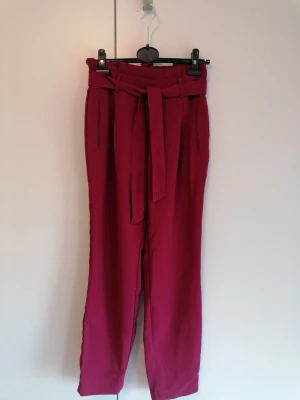 Höga rosa kostymbyxor med knytskärp - Snygga höga kostymbyxor i stark rosa färg med tillhörande knytskärp i midjan. Byxorna har raka ben och fickor framtill. Perfekta för dig som vill sticka ut med färg och stil. Materialet känns mjukt och följsamt. Bra skick men har blivit lite noppig på baksidan. 