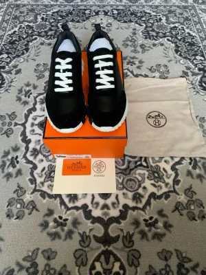 Svarta H E R M E S sneakers med vit sula - Snygga svarta sneakers med vit sula. Materialet ser ut att vara en mix av textil och mocka för en exklusiv känsla. Perfekta för dig som gillar stilrena och lyxiga detaljer.