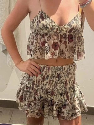 Blommigt tvådelat set med volanger - Säljer ett superfint tvådelat set med blommigt mönster i lila och svart på ljus botten. Toppen har smala axelband och volanger, kjolen är kort med flera volanglager. Perfekt för sommarens varma dagar. Materialet känns lätt och luftigt.