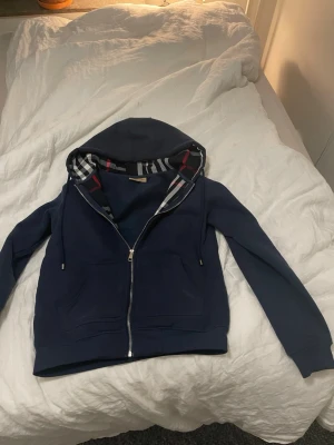 Marinblå zip-hoodie från Burberry - Snygg marinblå hoodie från Burberry med dragkedja framtill och klassiskt rutigt foder i huvan. Hoodien har fickor på magen och justerbara snören i huvan. Perfekt för dig som gillar stilrena detaljer och exklusiva märken.