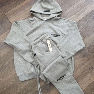 Grå Essentials hoodie med tryck - Säljer en grå hoodie från Essentials med svart logotyp-tryck på bröstet. Hoodien har en klassisk passform, ribbade muddar och en stor huva. Perfekt för dig som gillar en enkel och stilren look. Allt I bilden ingår. Helt nytt. Fraktar inom 24 timmar