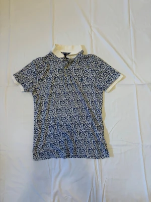 Blommig pikétopp från Polo Ralph Lauren - Säljer en vit och blå blommig pikétopp från Polo Ralph Lauren. Toppen har korta ärmar, klassisk krage och knappar framtill. Materialet är mjukt och skönt, perfekt för vår och sommar. Snygg och tidlös design med diskret logga på bröstet.