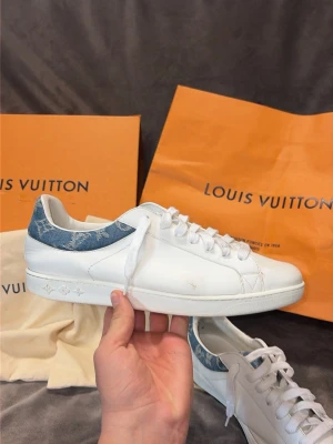 Louis Vuitton vita sneakers med denimdetalj - Säljer ett par vita Louis Vuitton sneakers i läder med blå denimdetalj och monogram runt hälen. Klassisk låg modell med vita skosnören och diskret präglad logga på sidan av sulan. Kommer med originalkartong och dustbags.