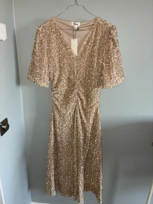 Champagnefärgad paljettklänning - Glittrig champagnefärgad klänning med v-ringning och korta ärmar. Klänningen är täckt av paljetter och har en smickrande rynkning framtill. Perfekt för dig som vill sticka ut med en elegant look.