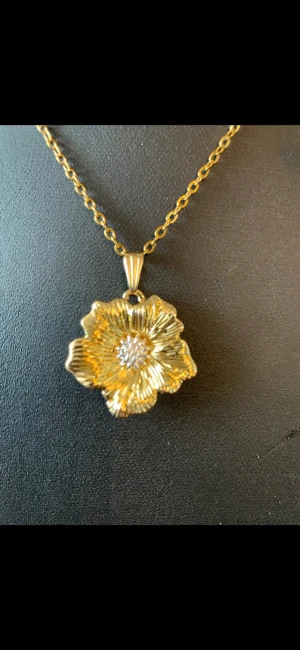 Guldigt halsband med blomma - Snyggt halsband i guldfärg med ett stort blomformat hänge. Hänget har detaljerade blad och en glittrande mitt, vilket ger en lyxig och trendig känsla. Perfekt accessoar för att lyfta din outfit med en unik touch. Kedjan är också guldfärgad och har klassisk länkdesign.