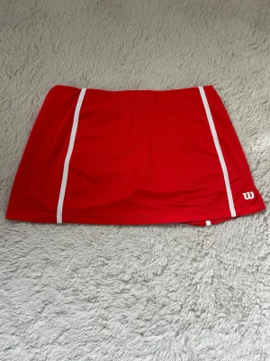 Röd tenniskjol från Wilson - Sportig röd tenniskjol från Wilson med vita ränder längs sidorna och diskret logga nere till höger. Tillverkad i ett lätt och stretchigt material som passar perfekt för intensiva matcher på banan.
