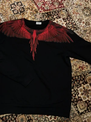 Marcelo Burlon sweatshirt  - Svart hoodie från Marcelo Burlon med ikoniskt rött fågeltryck över axlar och bröst. Tröjan har rund halsringning och ribbade muddar. Materialet känns mjukt och bekvämt, perfekt för en streetwear-look. Snygg detalj med vingarna som fortsätter på ryggen. Lite oversized passar S-M