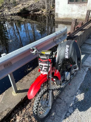 Röd crossmotorcykel med hjälm - Säljer en röd crossmotorcykel med svart och vit grafik, utrustad med Pro Taper-styre och nummerplåt med siffran 92. Medföljer svart Raven-hjälm. Perfekt för dig som gillar offroad och äventyr på två hjul.