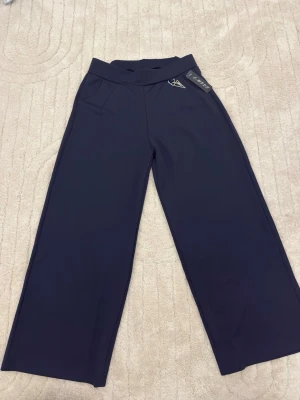 Vida mörkblå byxor  - Säljer ett par mörkblåa byxor med vida ben och hög midja. Byxorna har en snygg detalj med metallogo vid fickan och är tillverkade i ett mjukt, stretchigt material. Perfekta för en chill och trendig look.