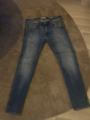Blå slim fit jeans från Jack & Jones - Snygga blå slim fit jeans från Replay/anbass med slitningar och lätt tvättad look. Klassisk femficksmodell med dragkedjegylf och läderpatch bak i midjan. Perfekta för en avslappnad och trendig stil. Storlek 31/30. Lite hål vid skrevet men det är jätte litet. Hör av er vid frågor pris går att diskutera 