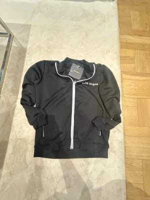 Svart zip hoodie från Palm Angels - Palm Angels svart zip-tröja med vit dragkedja och vit logotyp på bröstet. Tröjan har ribbade muddar och två fickor med dragkedja. Materialet är bomul perfekt för en cool streetwear-look. Snabb shipping 