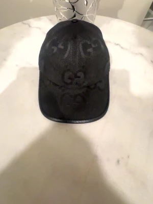 Svart keps från Gucci med GG-mönster - Snygg svart keps från Gucci med det klassiska GG-mönstret över hela kepsen. Kepsen har böjd skärm och är tillverkad i ett tygmaterial som ger en exklusiv känsla. Perfekt accessoar för att lyfta din streetwear-look.