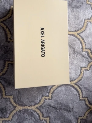 Svarta sneakers från Axel Arigato - Snygga svarta sneakers från Axel Arigato med vit sula och elastiskt band över vristen. Skorna är i mocka och har diskret guldfärgad logga på sidan. Perfekta för dig som gillar stilrena och minimalistiska sneakers.