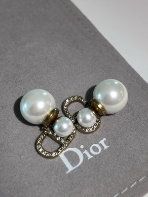 Dior pärlörhängen med strassdetaljer - Snygga örhängen från Dior med vita pärlor i två storlekar och guldfärgade detaljer. Örhängena har glittrande strass på Dior-loggan som ger extra lyxig känsla. Perfekt accessoar för dig som gillar elegant och trendig stil.Without box.