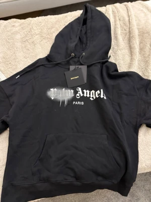 Svart Palm Angels hoodie med tryck - Svart hoodie från Palm Angels med stor huva och dragsko. Framsidan har ett vitt tryck med texten 'Palm Angels Paris' och en cool spray paint-effekt. Stor känguruficka framtill. Perfekt för en streetwear-look. Helt oanvänd pga liten storlek 