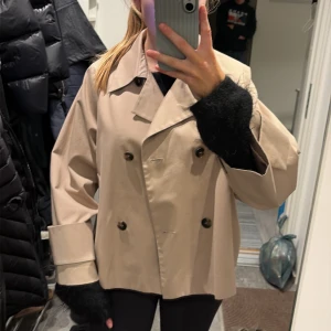 Beige kort trenchcoat med stora slag - Snygg beige trenchcoat med dubbelknäppning och stora slag. Jackan är kortare i modellen och har breda ärmslut samt fickor i sidorna. Perfekt för dig som gillar en stilren och trendig look. Passar till många outfits och är lätt att matcha.