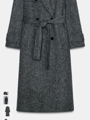 Grå lång ullrock med knytskärp - Stilren grå lång rock i ullmix med dubbelknäppning och knytskärp i midjan. Rocken har breda slag, två stora fickor framtill och detaljer vid ärmsluten. Perfekt för kyliga dagar och ger en klassisk look.