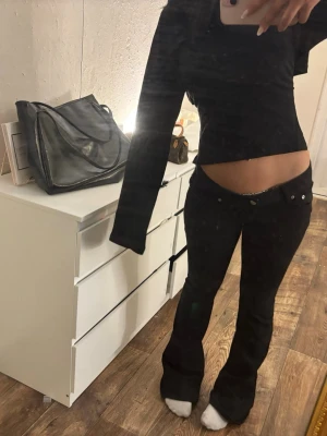 Svarta low waist bootcut jeans - Säljer ett par svarta low waist jeans med bootcut passform. Jeansen har silverknappar och detaljer vid fickorna. Sitter tight över höfterna och har utsvängda ben. Perfekta för dig som gillar en trendig och edgy stil.