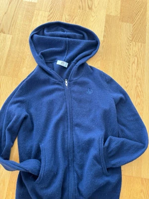 Mörkblå kashmir hoodie från Bvaldi - Mysig mörkblå hoodie från Bvaldi med dragkedja framtill och två fickor. Tillverkad i mjukt material och har en broderad logga på bröstet. Perfekt för chill dagar eller när du vill ha något bekvämt och stilrent. Klassisk huva för extra komfort.