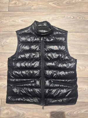 Svart dunväst från Moncler - Snygg svart dunväst från Moncler med hög krage och två dragkedjeförsedda fickor framtill. Västen har ett quiltat mönster och Moncler-logga på bröstet. Perfekt för lager-på-lager och håller dig varm med stil. Den är i storlek 6 vilket motsvarar en större xl