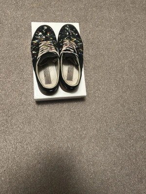 Maison Margiela - Unika svarta sneakers från Maison Margiela med färgstänk i flera färger över hela ovandelen. Skorna har vita snören och en klassisk låg siluett. Tillverkade i läder med en cool och kreativ design som sticker ut i mängden.