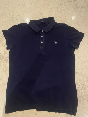 Mörkblå pikétröja från Gant - Klassisk mörkblå pikétröja från Gant med korta ärmar och krage. Tröjan har fyra knappar framtill och en liten broderad logga på bröstet. Tillverkad i mjukt bomullsmaterial som känns bekvämt mot huden.