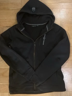 Svart hoodie från Sail Racing - Svart hoodie från Sail Racing med dragkedja framtill och logga på bröstet. Tröjan har huva med broderad detalj och vita kontrastsömmar vid halsen. Tillverkad i mjukt material som känns skönt mot huden. Perfekt för en avslappnad och sportig stil.